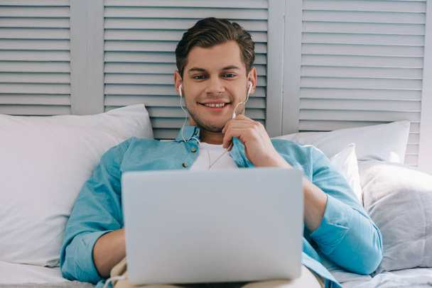 Young man in earbuds using laptop while lying on bed - Foto, Imagem