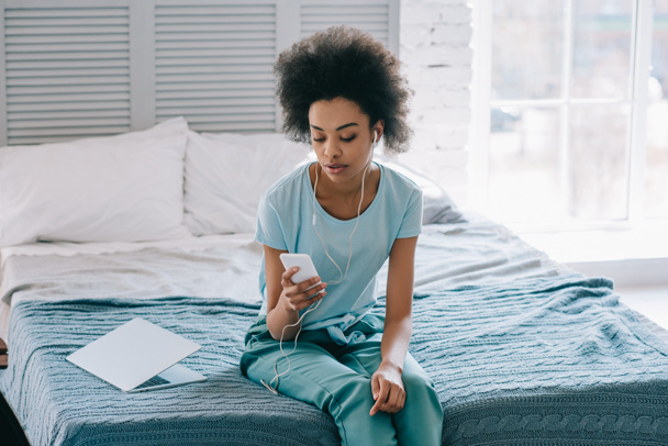 African american woman sitting on bed and listening to music on phone - Photo, Image