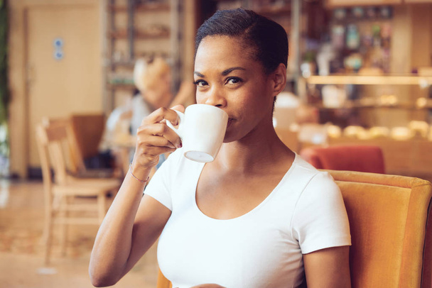nézi a kamera vesz egy kortyot a bögre kávét egy modern kávézóban közben a gyönyörű afrikai nő - Fotó, kép