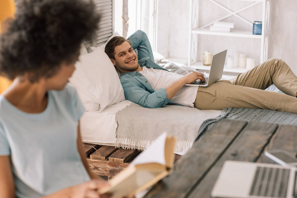 African american woman reading book by man lying on bed with laptop - Photo, Image
