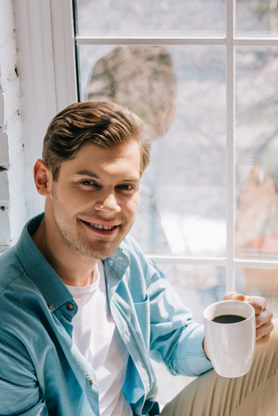 Un homme souriant tenant une tasse de café assis sur le rebord de la fenêtre
 - Photo, image