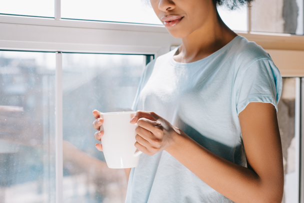 Nahaufnahme der Kaffeetasse in den Händen eines afrikanisch-amerikanischen Mädchens am Fenster - Foto, Bild