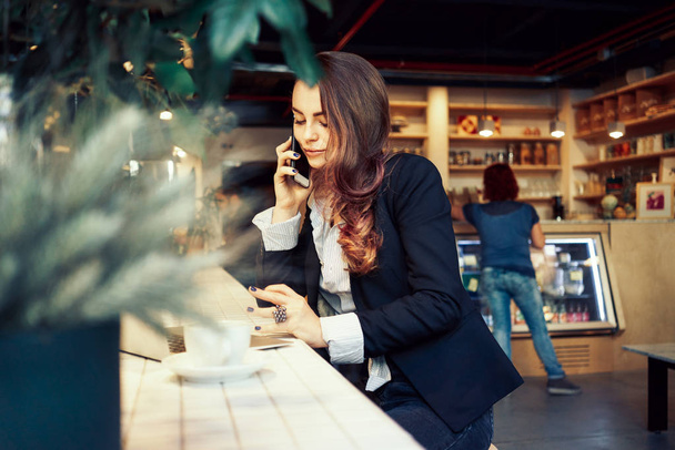 Mulher de negócios moderna atraente fazendo uma chamada de negócios e usando um laptop pessoal enquanto sentado em uma cafeteria
 - Foto, Imagem