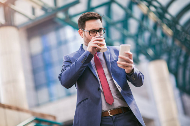 Бізнесмен використовує мобільний телефон і п'є каву за межами офісної будівлі на задньому плані
.  - Фото, зображення