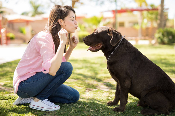Wesoły młoda kobieta latin dmuchanie pocałunek czekoladowy labrador w odkryty park. - Zdjęcie, obraz