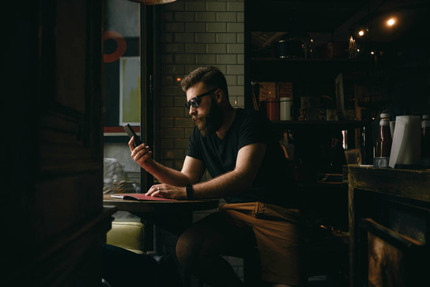 Handsome bearded man wearing sunglasses looking at the screen of smartphone at coffee shop - Фото, изображение