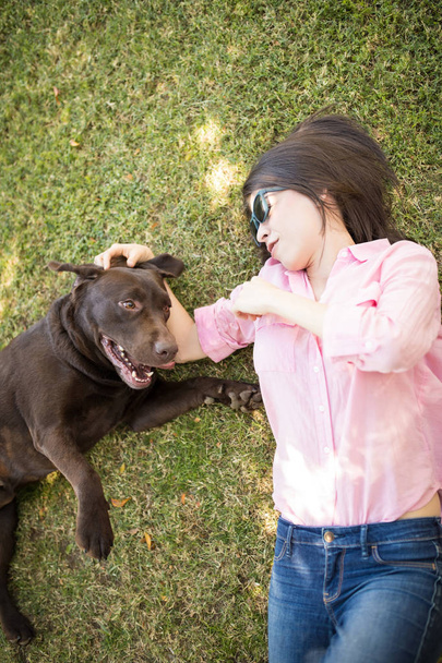 Side view of young woman lying on green grass petting dog lying next to her. - Photo, Image