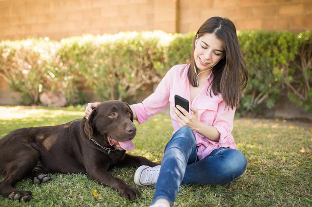 Enjoying woman sitting in park with earphones and smartphone besides pet dog - Photo, Image