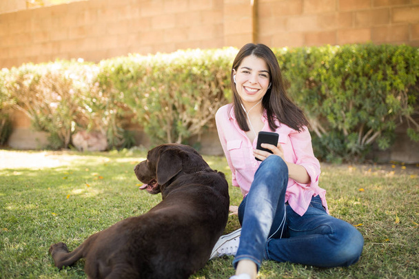 Cheerful young woman holding smartphone with earplugs sitting besides chocolate labrador in park. - Photo, Image