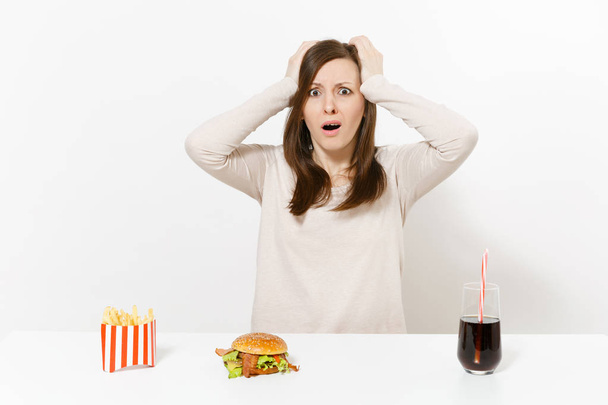 Shocked woman put hands on head at table with burger french fries cola in glass bottle isolated on white background. Proper nutrition or American classic fast food. Advertising area with copy space. - Photo, Image