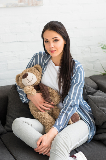 portrait of smiling woman with teddy bear resting on sofa at home - Photo, Image