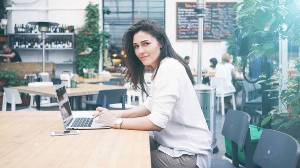 Young beautiful woman with long dark hair working with laptop while sitting on a terrace of a street coffee shop  - Photo, Image
