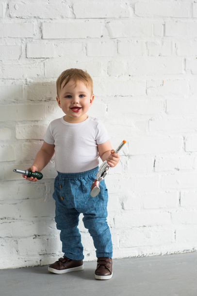 mutlu küçük oğlu beyaz tuğla duvarın ayakta oyuncaklar ile - Fotoğraf, Görsel