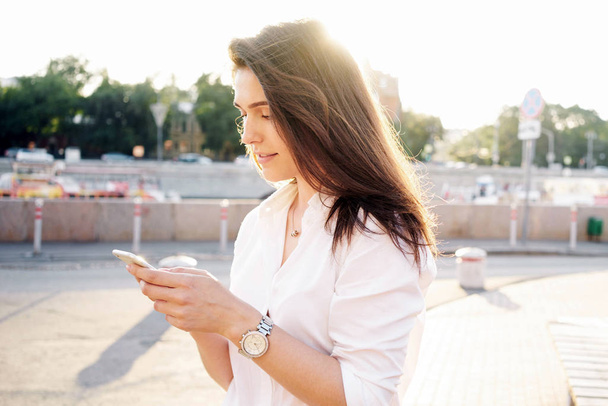 Beautiful young woman holding a smartphone in the hands while standing in a street on a sunny day  - Foto, Imagem