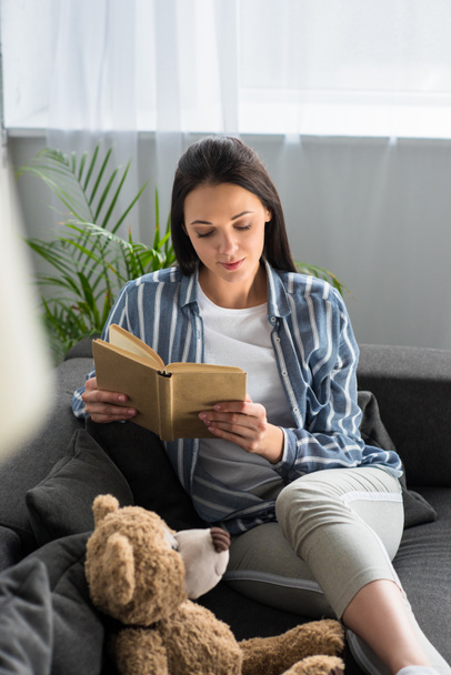young woman reading book on sofa with teddy bear at home - Foto, imagen