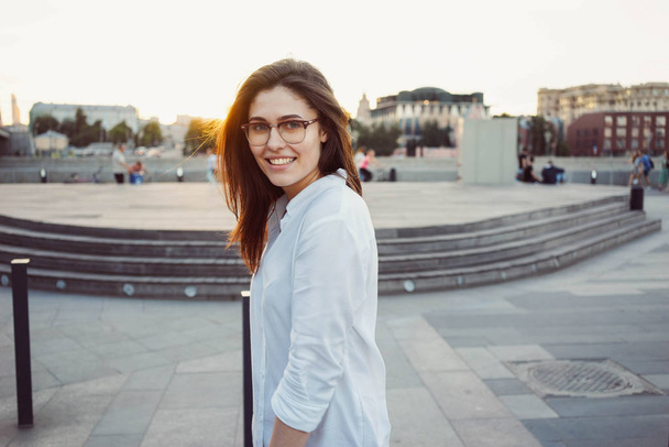 A cropped photo of a beautiful brunette woman in the glasses wearing white shirt posing on a city park background - Fotografie, Obrázek