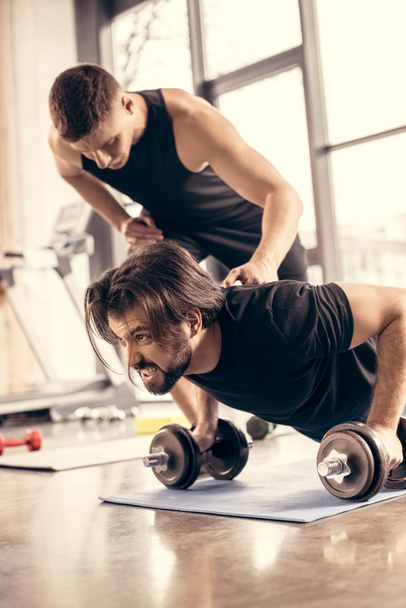 trainer pushing sportsman doing push ups on dumbbells in gym - Photo, Image
