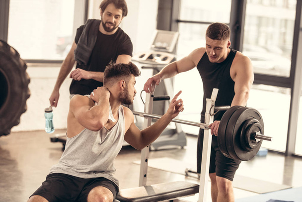 handsome sportsman having pain in neck and talking with friends in gym - Photo, Image