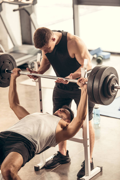 мышечный тренер помогает спортсмену поднимать штангу с тяжелыми грузами в тренажерном зале
 - Фото, изображение