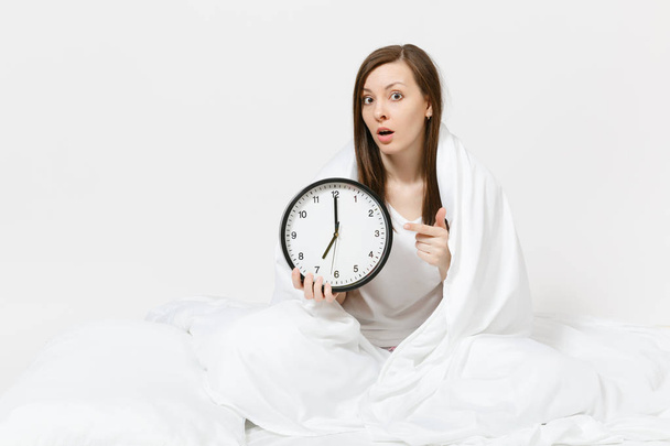 Young woman sitting in bed with round clock, white sheet, pillow, wrapping in blanket on white background. Beauty female wake up early in morning, spending time in room. Rest, relax, good mood concept - Fotoğraf, Görsel