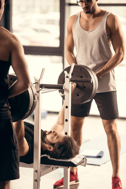 sportsmen helping friend lifting barbell in gym - Photo, Image