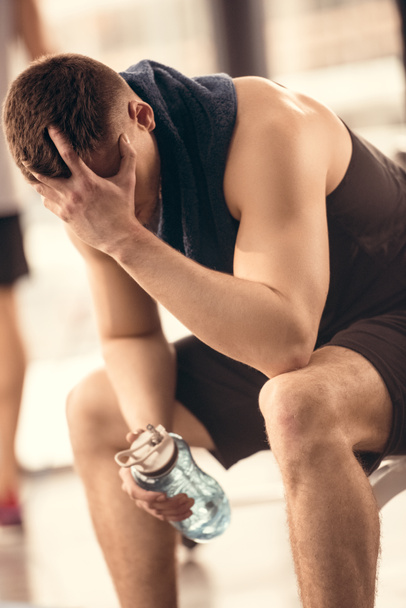 κουρασμένος αθλητή κρατώντας μπουκάλι νερό και αναπαύεται στο γυμναστήριο - Φωτογραφία, εικόνα