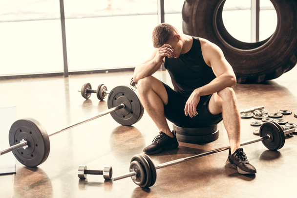 upset young sportsman sitting with dumbbells and barbells in gym - Photo, Image