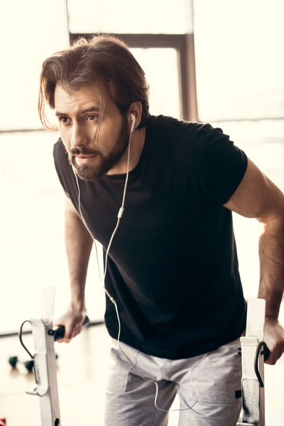 muscular young man in earphones exercising on bars and looking away in gym  - Zdjęcie, obraz