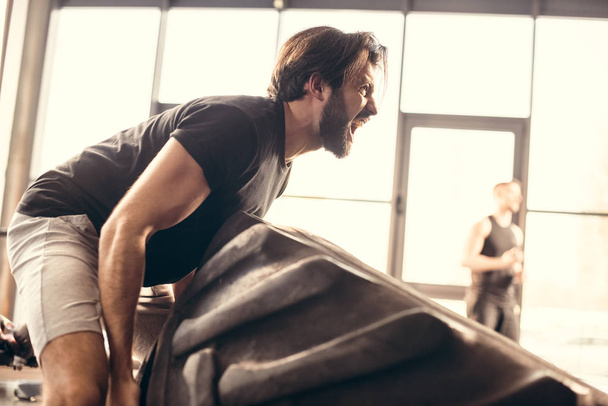 side view of athletic man lifting tire and screaming in gym  - Fotografie, Obrázek