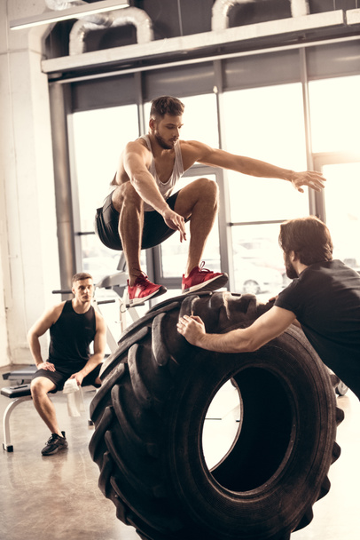 спортивний молодий чоловік стрибає на шині під час тренувань з друзями в спортзалі
 - Фото, зображення