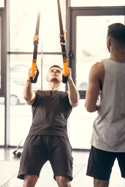 oříznuté záběr sportovní člověka při pohledu na svalnatého muže školení s popruhy suspenze v tělocvičně - Fotografie, Obrázek