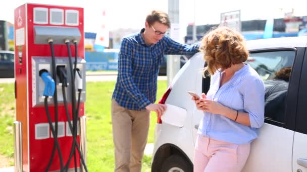 Smiling young couple on charging station for electric cars. Man charging car and woman standing with smartphone - Video