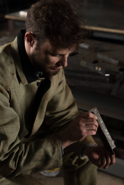 vista de ángulo alto del joven trabajador masculino sentado y mirando la parte metálica en la fábrica
 - Foto, Imagen