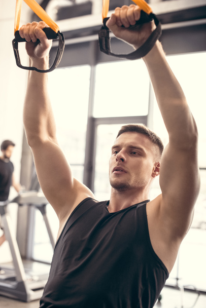 handsome athletic young man exercising with fitness straps and looking away in gym - Foto, Bild