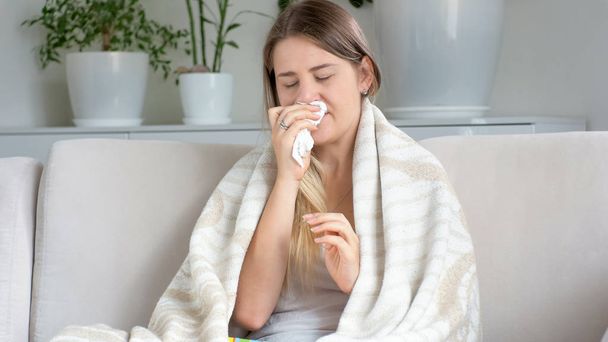Portrait of sick young woman covered in blanket sneezing in tissues - Φωτογραφία, εικόνα