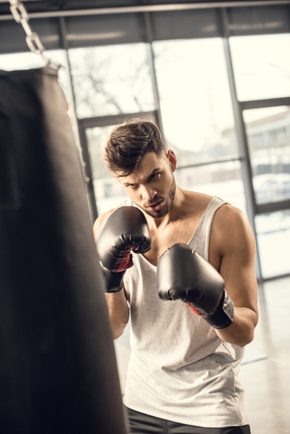 jeune sportif concentré dans des gants de boxe regardant sac de boxe dans la salle de gym
 - Photo, image