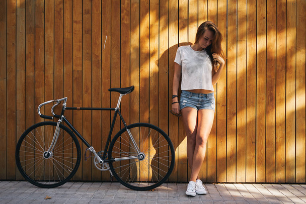 Young sporty woman wearing white blank t-shirt and jeans shorts standing with bicycle on the wooden background - Fotó, kép