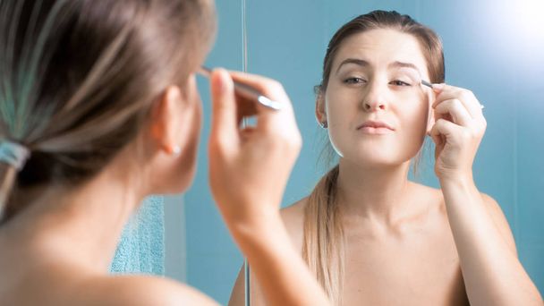 Portrait of beautiful young woman using tweezers at mirror - Photo, image