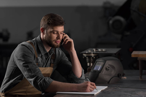 trabajador masculino escribiendo en libro de texto y hablando en smartphone
  - Foto, Imagen