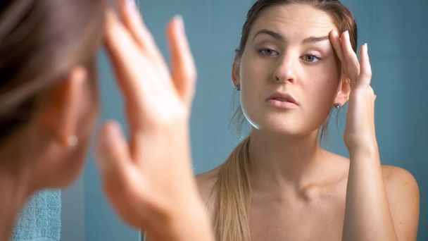 Портрет брюнетки, осматривающей свою кожу у зеркала в ванной комнате
 - Фото, изображение