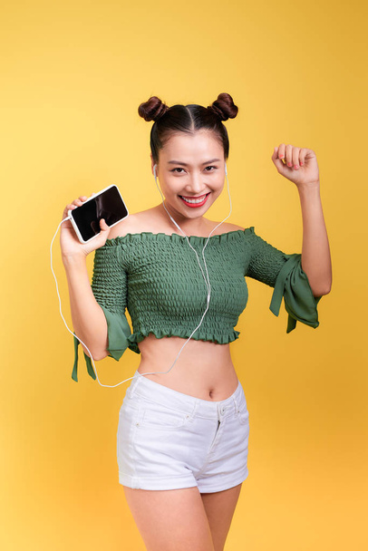 Fashion smiling asian woman listening to music in earphones  over yellow background - Foto, immagini