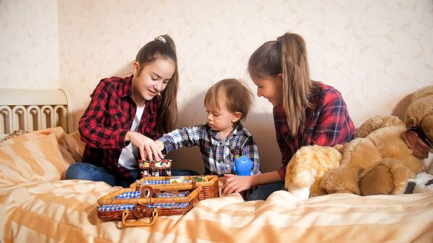 Две девочки-подростки играют со своим младшим братом с игрушками на кровати
 - Фото, изображение