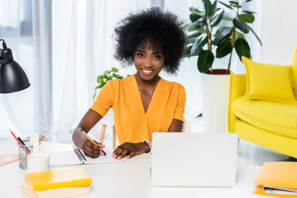 portrait of smiling african american freelancer at workplace with laptop and notebook at home - Фото, зображення