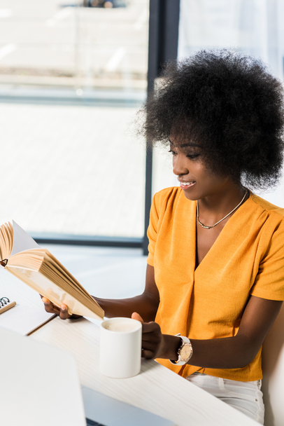 усміхнений афроамериканський американець з чашкою кави читає книгу за столом в домашньому офісі
 - Фото, зображення