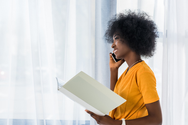 side view of african american freelancer with folder talking on smartphone at home - Photo, Image