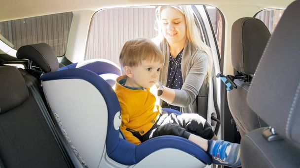 Beautiful smiling mother seating her toddler son in safety seat - Photo, Image