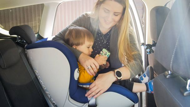 Portrait de jeune mère mettant son fils tout-petit dans le siège de sécurité de voiture
 - Photo, image