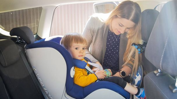 Portrait of young mother fastening belts and bucle of car children safety seat - 写真・画像