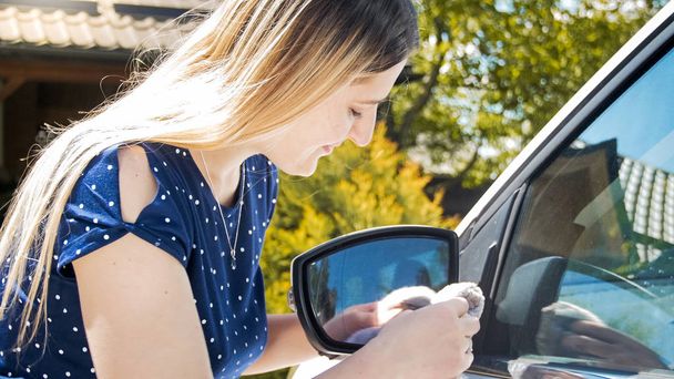 junge lächelnde Frau wischt mit Tuch Seitenspiegel ihres Autos ab - Foto, Bild