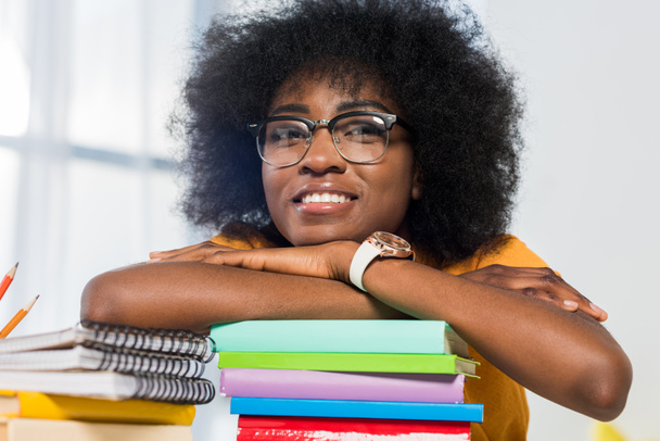 sonriente afroamericano freelancer en anteojos apoyados en un montón de cuadernos en el lugar de trabajo en la oficina en casa
 - Foto, Imagen